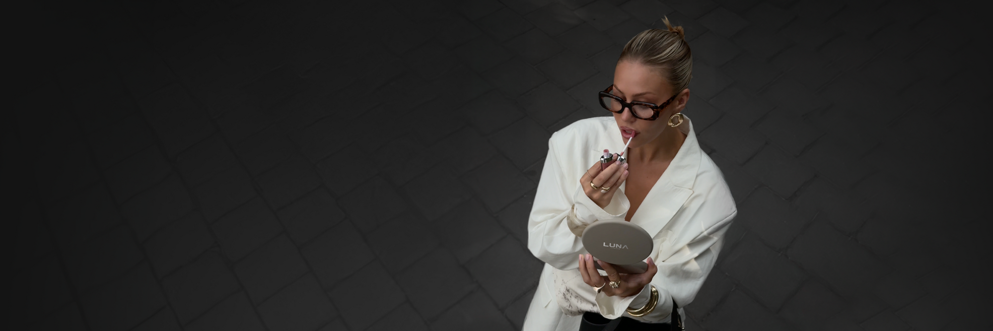 A woman with blonde hair in a bun and black glasses, wearing a white robe, holds a small round makeup compact while applying makeup. She is photographed from a high-angle perspective against a dark gray tiled background.