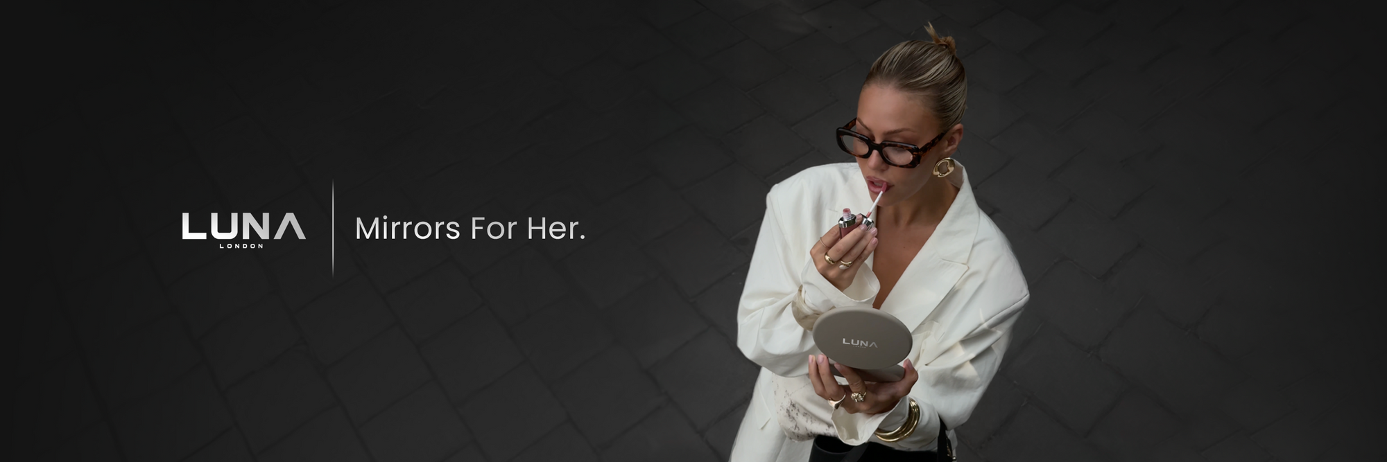 A woman in a white robe looks into a round LUNA Paris compact mirror while applying lip product. The scene is set against a dark tiled background with the text 'LUNA Paris Mirrors For Her.